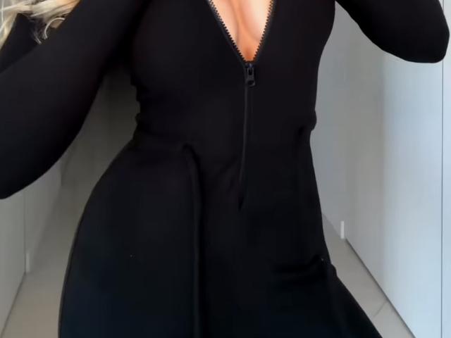 A MargoFun está usando um vestido preto com zíper, que revela seus seios através do decote em V, com o bumbum ligeiramente levantado, cabelo loiro parcialmente visível, em um espaço estreito com paredes cinzas..