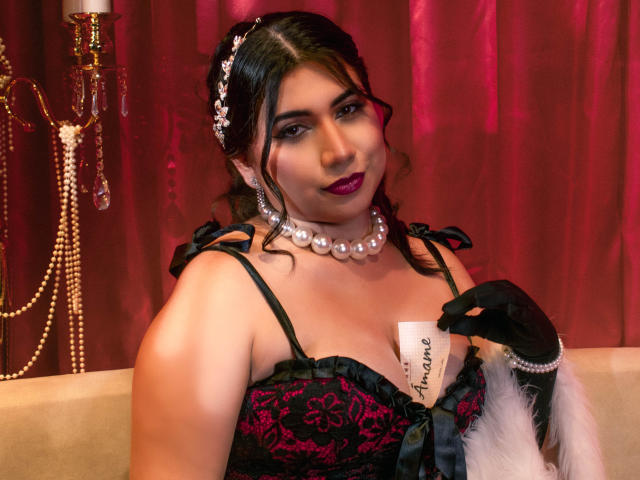 BabeCloe with dark hair in a floral headpiece, wearing a black and red lace corset, pearl necklace, and black gloves. She holds a white tag with "Santiago" in cursive. Her makeup includes dark purple lipstick. A white fur stole is draped over her right arm. Red curtain backdrop. Warm, intimate lighting. Detailed lace patterns and pearl texture. BabeCloe's confident, slightly smirking expression. Rich colors and textures. Glamorous, vintage-inspired look.