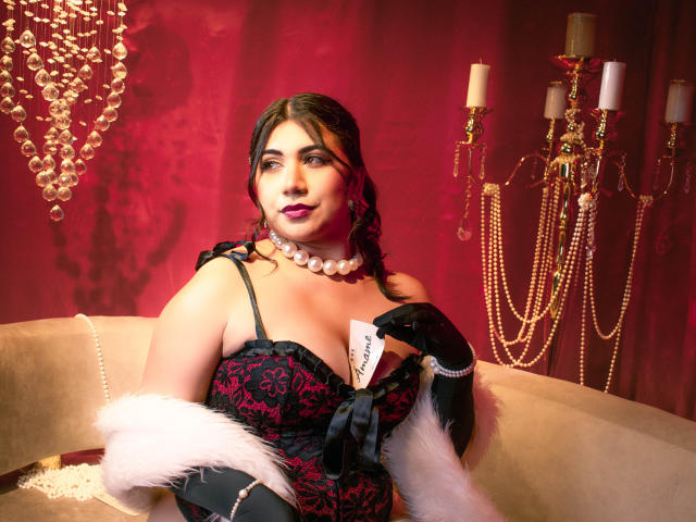 BabeCloe is in a rich red room, wearing a black and red lace corset with a low neckline, showing a tag that reads "SILK." She has dark wavy hair, a pearl necklace, and black gloves with white fur. She's holding a pearl necklace in her right hand. A gold candelabrum with white candles is in the background. The lighting is warm and highlights her skin and corset. She has a confident expression.