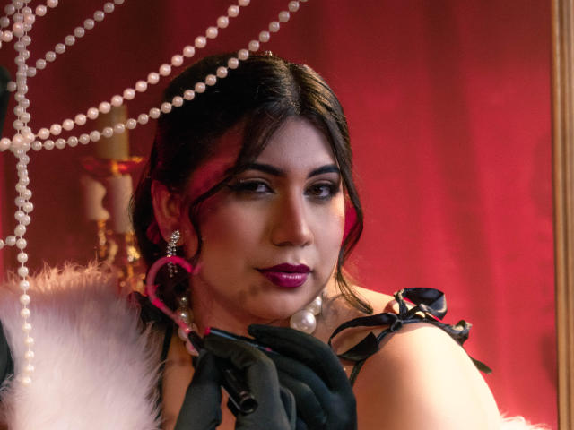 BabeCloe with dark hair, pink highlights, purple lipstick, pearl necklace, black gloves, and pink earrings, looking smoky-eyed, holding black straps, under white pearls and red lighting.
