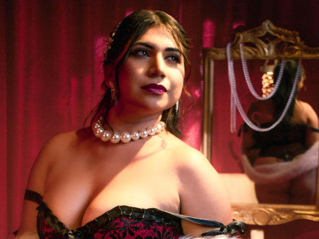 BabeCloe with large boobs, wearing a black and pink lace corset, pearl necklace, and dark makeup, looking in a mirror with pearl jewelry in the reflection.