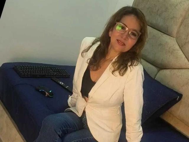MargarethRouss, with wavy brown hair and glasses, sits on a blue bed, wearing a white blazer over a black top, and blue jeans, with a remote and keyboard nearby.