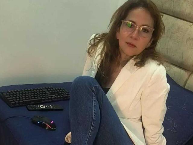 MargarethRouss is sitting on a bed with blue sheets, wearing glasses, a white jacket, and blue jeans, with a keyboard and mouse in front of her.