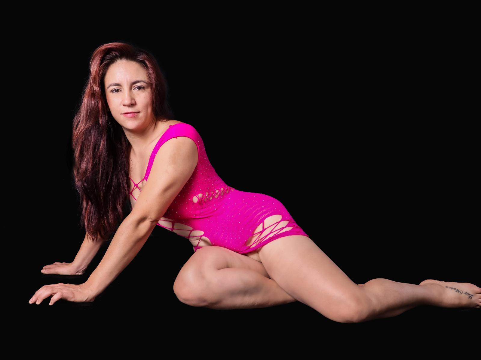 SofiaMartinez69, with red hair, pink mesh dress, and visible pussy through cutouts, is on all fours, legs spread, black background, smiling at the camera.