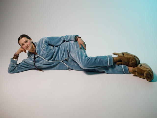 TayaFlavy lies on a white floor, wearing a blue, textured, zip-up jumpsuit and tan boots. She rests her head on her right hand, smiling slightly. Her left arm is bent, supporting her upper body. TayaFlavy has a light brown face, dark brown hair in a braid, and wears a black wristwatch on her right wrist. The image has a soft, blue gradient in the top right corner. The composition is simple, focusing on TayaFlavy's relaxed, casual pose.