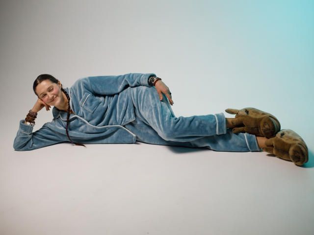 TayaFlavy lies on a flat surface, wearing a blue denim jumpsuit with white stripes. She has dark hair in a ponytail, smiling with her head resting on her right hand. Brown boots with visible wear sit on her feet. She wears multiple brown bracelets on her left wrist. The composition is simple, with a light gradient from blue to white in the background. The image has a relaxed, casual vibe. Her relaxed posture and casual attire suggest a laid-back, everyday style.