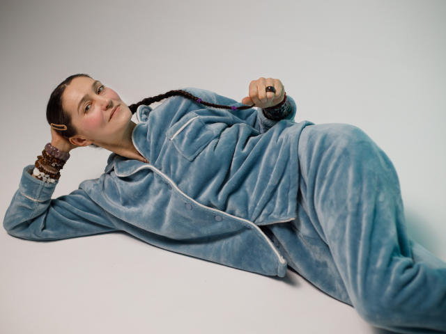 TayaFlavy is lying on a flat surface in a relaxed pose. She has light skin, brown eyes, and long, braided hair. She's wearing a blue, velvety zip-up jacket and matching pants. Her left hand, adorned with brown beaded bracelets, rests on her head while her right hand holds a black ring. The light gray background is plain, focusing all attention on TayaFlavy and her casual, comfortable outfit. Her expression is neutral, and she looks slightly to the side. The composition is simple, emphasizing her relaxed, laid-back demeanor.