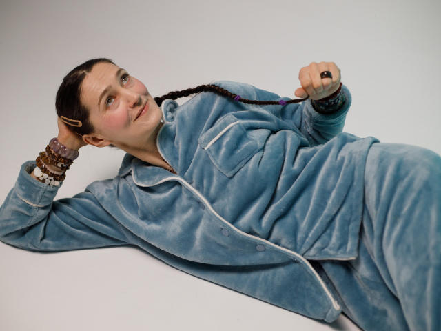 TayaFlavy lies on a white surface, wearing a light blue, velvety zip-up outfit. She has a fair complexion and brown hair in a single braid. She holds a black ring in her right hand while her left hand rests on her head. She wears multiple brown and purple bracelets. Her expression is neutral, and she gazes slightly to the left. The texture of the velvety outfit contrasts with her smooth skin. The composition is simple, with TayaFlavy positioned diagonally across the frame.