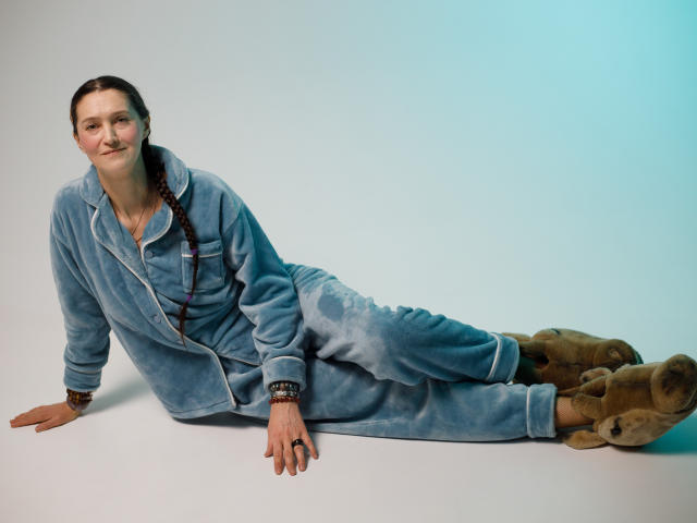 TayaFlavy is lying on a flat surface in a relaxed pose, wearing a light blue, velvety jumpsuit with rolled-up sleeves and cuffs. She has a thin mustache and is looking at the camera with a slight smile. Her dark brown hair is in a braid over her right shoulder. She has a black bracelet on her left wrist and a ring on her right hand. Her feet are covered in brown, furry slippers. The image has a cool, blue gradient in the background.