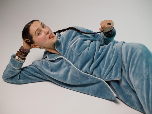 TayaFlavy lies on her side in a relaxed pose, wearing a light blue, velvety, button-up suit. She has a braided brown ponytail and a slightly open mouth with a playful expression. Her right hand rests on her head, while her left hand holds a thin string. She wears multiple brown and purple bracelets on her left wrist and a dark ring on her right index finger. The image focuses on her casual, relaxed posture and the texture of the velvety suit.