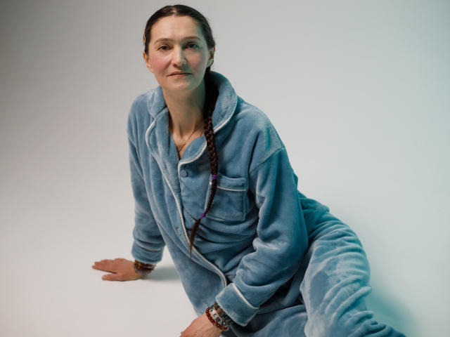 TayaFlavy sits on a white surface, leaning back on her right hand. She wears a light blue, fluffy, zip-up onesie with white trim. Her brown hair is in a single braid, hanging over her left shoulder. She has a fair complexion, subtle freckles, and a neutral expression. Her left hand rests on the floor, and she wears a bracelet with small beads on her right wrist. The image has a soft, natural light, emphasizing the textures of the onesie and her hair.