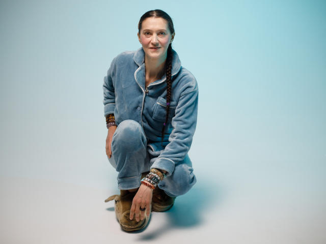 TayaFlavy kneels on a light blue studio floor, wearing a gray zip-up fleece with a front pocket and light blue pants. She has brown boots and multiple beaded bracelets on both wrists. Her brown hair is in a single braid over her right shoulder. She's smiling slightly, looking directly at the camera. The image has a clean, minimalistic style with a bright, even light.