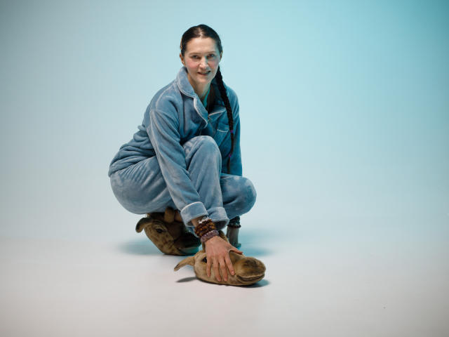 TayaFlavy, squatting, wearing a blue jumpsuit, brown boots, and a bracelet, with a slight smile, hands on boots, against a plain background.