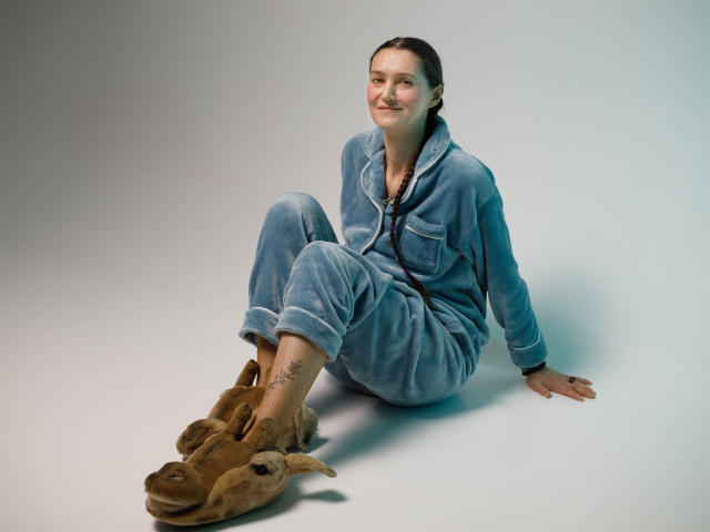TayaFlavy sits on a light grey floor, wearing a blue, fuzzy, zip-up onesie with rolled-up cuffs. She has a small tattoo on her left ankle. Her brown hair is pulled back in a simple style. She's smiling slightly, looking to the right. Brown, plush slippers with animal-like features are in front of her. The lighting is even, highlighting her relaxed posture and casual attire. The image is clear with a soft focus.