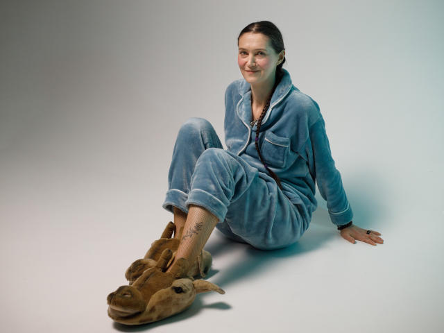 TayaFlavy sitting on floor, wearing blue denim pajamas, brown sheep slippers, smiling, tattoo on right ankle, brown hair in ponytail, light skin, relaxed pose.