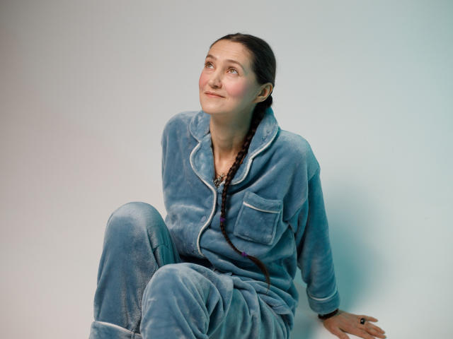 TayaFlavy, a woman with fair skin and dark braids, sits against a light gray background. She wears a blue velvet pajama set with white trim, looking slightly to the right with a gentle smile. Her left hand rests on the floor while her right arm is relaxed. Soft light highlights her cheekbones and the texture of the velvet. Her expression is calm and slightly curious.