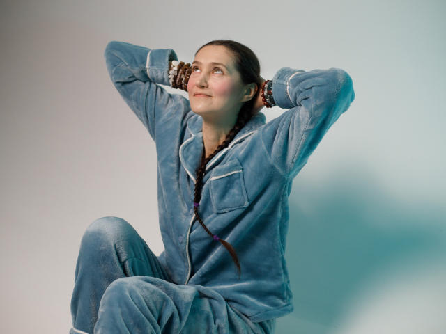 TayaFlavy sits with one knee up, hands behind her head, looking slightly to the right. She wears a blue velvet pajama set with white piping, her brown hair in a single braid. Her skin is fair, and she has a subtle, confident smile. The light is soft, creating a shadow on the right. Her relaxed posture contrasts with the formal, buttoned pajamas. The image uses cool tones, with a focus on texture and TayaFlavy's casual, yet poised, demeanor. The composition is balanced, with TayaFlavy centered.