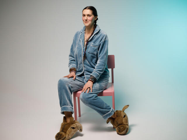 TayaFlavy sits on a pink chair, wearing a blue, oversized denim jumpsuit with rolled cuffs and brown beaded bracelets. She has light skin, brown hair in a low ponytail, and a relaxed expression. She's wearing tan, furry slippers shaped like small animals. The image has a clean, minimalist composition with a subtle gradient from grey to blue in the background. The chair is simple with a pink frame. TayaFlavy's casual, relaxed posture contrasts with the structured denim outfit.