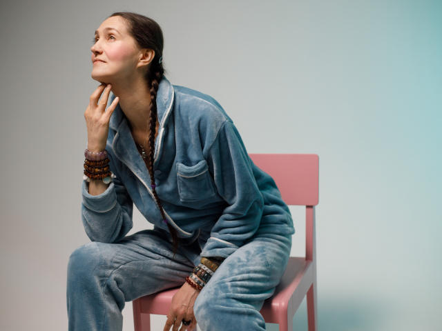 TayaFlavy with braided hair, wearing a blue fuzzy onesie, sits on a pink chair, looking up with a thoughtful expression, hand on cheek, wearing multiple brown bracelets.
