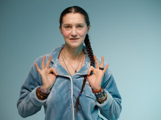 TayaFlavy with braids, blue fuzzy hoodie, hands up, showing fingers, smiling, wearing bracelets and necklace, light skin, medium-sized tits, soft lighting, calm expression.