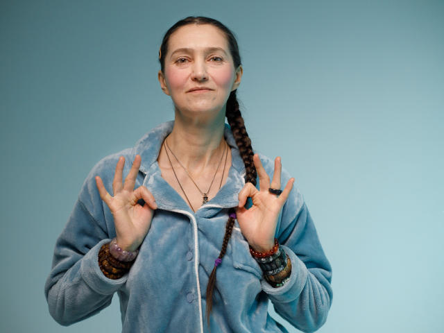 TayaFlavy with braided hair, wearing a blue fleece jacket, holding hands up, smiling slightly, wearing multiple bracelets and a necklace, against a plain background.