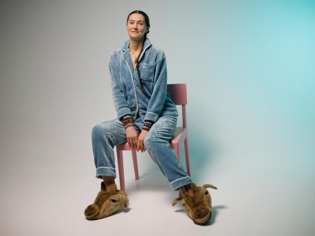 TayaFlavy sitting on pink chair, wearing denim jumpsuit, rolled cuffs, brown chunky boots, smiling, hands on knees, light blue-green gradient background, light skin, brown hair, relaxed.