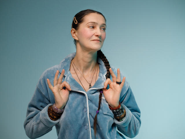 TayaFlavy has a light skin tone and a slender build. She's wearing a blue, fluffy zip-up jacket with white trim. Her dark brown hair is in a single braid over her right shoulder. She has small, slightly upturned nose and is looking to the right with a subtle, confident smile. She's making a double rock gesture with both hands, fingers spread. She has a thin necklace with a small cross and multiple colorful bracelets on her left wrist. A small hair clip is in her right braid. The image is sharp with even lighting.