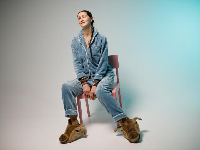 TayaFlavy sits on a red chair, leaning back slightly with a relaxed expression. She wears a blue denim overall, cuffed at the ankles, with brown suede boots. Her dark braids are pulled back, and she has multiple brown and black bracelets on her right wrist. The overall is loose-fitting, contrasting with the tightness of her boots. The light is soft, creating a subtle gradient from white to blue on the right. Her hands are resting on her knees, fingers loosely interlaced. The composition is centered, with TayaFlavy's relaxed posture adding a casual vibe.