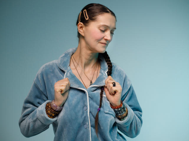 TayaFlavy stands against a solid blue background. She has fair skin, dark brown hair in a braid, and small hair clips. She wears a light blue, fuzzy hoodie with white buttons, and multiple bracelets on her right wrist. She holds the hoodie's edges with her fists, looking slightly to her right with a subtle smile. A black ring is on her right index finger. Her eyes are closed, and she has a light blush on her cheeks. The image is well-lit and focused.