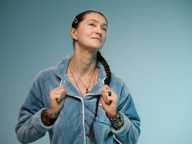 TayaFlavy stands against a solid blue background, wearing a blue fleece jacket with a slightly open front. She has a fair skin tone and dark brown hair in a braid. Her fists are clenched in front of her chest, showing a black ring on her right hand. She has a subtle smile, and multiple bracelets are on her right wrist. The image is sharp with a realistic texture on the jacket.