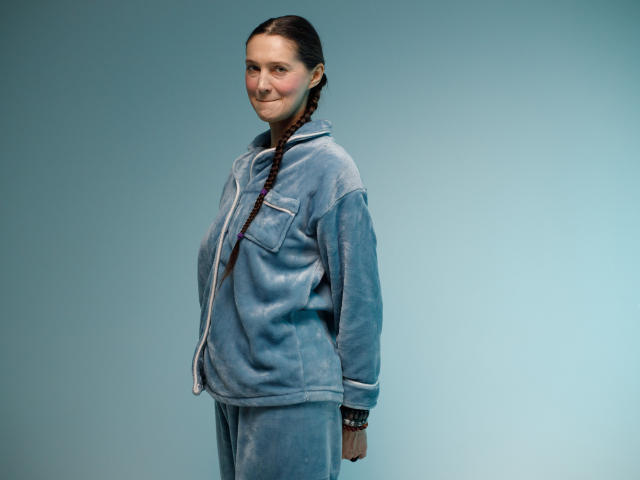 TayaFlavy, with braided brown hair, wears a light blue, velvety, oversized jacket and matching pants, standing, smiling slightly, hands behind her back, in a simple, blue-lit setting.