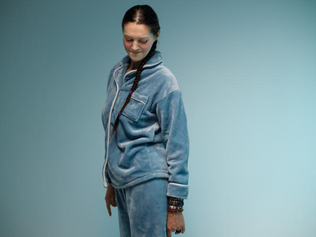 TayaFlavy stands with a slight forward lean, eyes downcast. She wears a light blue, velvety hoodie with white trim and matching pants. Her dark brown hair is in a single braid over her right shoulder. She has a light skin tone and a subtle, closed-lip smile. Her left wrist is adorned with multiple beaded bracelets. The background is a solid, light blue color. The image is a clear, well-lit photograph with a simple, focused composition.