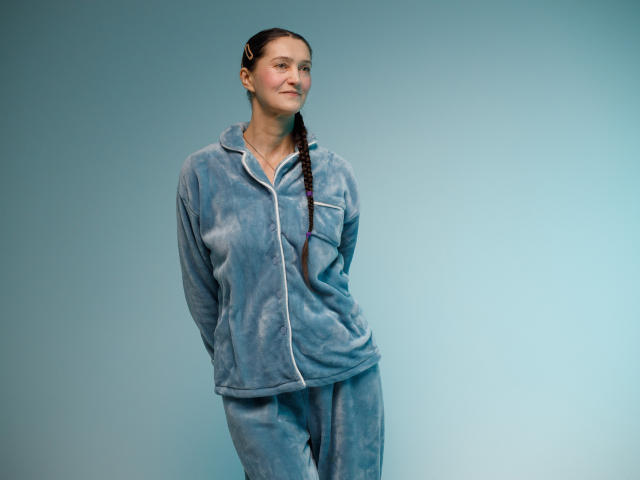 TayaFlavy stands against a plain blue background, wearing a light blue, fuzzy hoodie and matching pants. She has a slight smile, looking to the right, with her arms behind her back. Her brown hair is in a single braid that hangs to her side. The texture of the hoodie is soft and velvety, with a slightly worn look. The overall composition is simple and centered, focusing on TayaFlavy's relaxed, casual appearance.