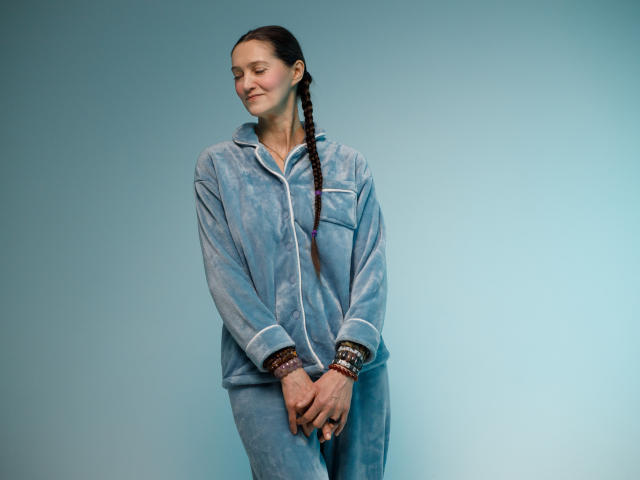 TayaFlavy stands against a light blue gradient background. She has fair skin, dark brown hair in a single braid, and a slight smile. She wears a loose, light blue, long-sleeve, zip-up hoodie with white trim and matching pants. Her hands are clasped in front of her, adorned with colorful beaded bracelets. Her relaxed posture and gentle smile create a calm, serene atmosphere. The image uses soft, even lighting to highlight her relaxed demeanor. The overall composition is simple and focused on TayaFlavy's relaxed, casual style.