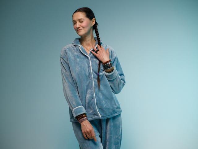 TayaFlavy with dark braids, wearing a blue tie-dye hoodie and matching pants, smiling softly, hand on chest, wrist bracelets, standing against a plain blue background.