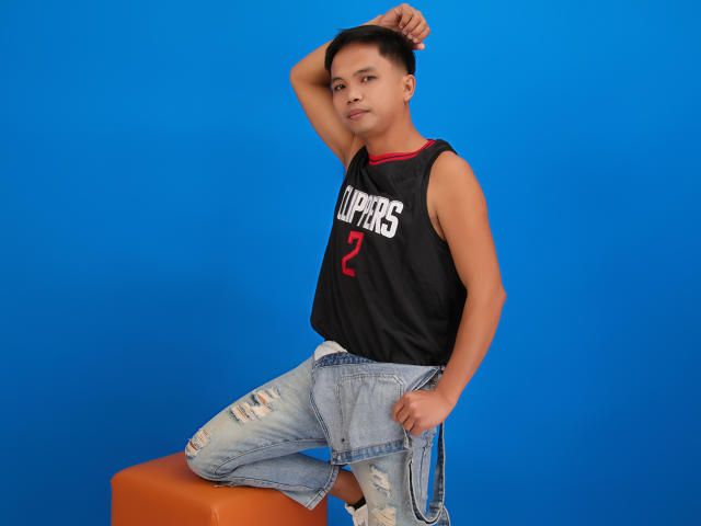 DerickLoganMan with short black hair, wearing a black "Supers" tank top and ripped jeans, sits on an orange stool, arm raised, against a bright blue background.