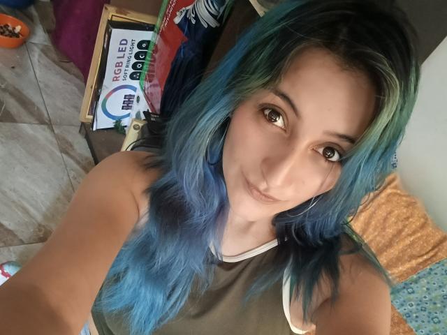 KarolLove6, a young woman with long, blue and green-highlighted hair, takes a selfie. She has light skin, brown eyes, and a small nose ring. She wears a brown sleeveless top with white trim. The background includes a cardboard box with "SCHOOL PACK" and a blue circular logo, and a bed with an orange pillow and blue patterned blanket. The image has a casual, homey feel. Her expression is neutral, and she looks directly at the camera. The photo is taken from a high angle.