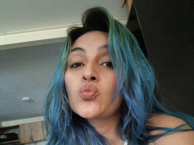 KarolLove6. has blue and green hair, pouting her lips. Her face is centered, with slightly parted, puckered lips. Her dark brown eyes look directly at the camera. She has a light brown skin tone. Her hair is messy, with blue and green highlights. She's wearing a white top. The background is blurry, showing a gray wall and part of a ceiling. The image is a close-up selfie. The lighting is natural.