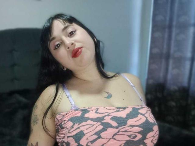 CrystalxSweet with black hair, pink floral top, and tattoos, pouting with red lips, showing her small tits, and a crescent moon tattoo on her chest.
