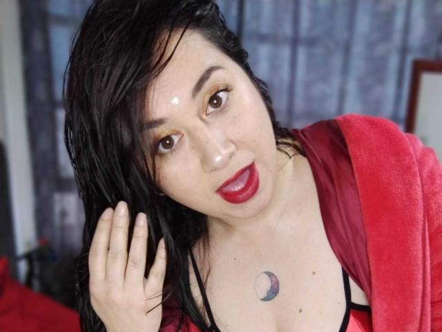 CrystalxSweet with wet black hair, red lipstick, and a crescent moon tattoo on her chest, wearing a red robe, touching her head with a slightly surprised expression.