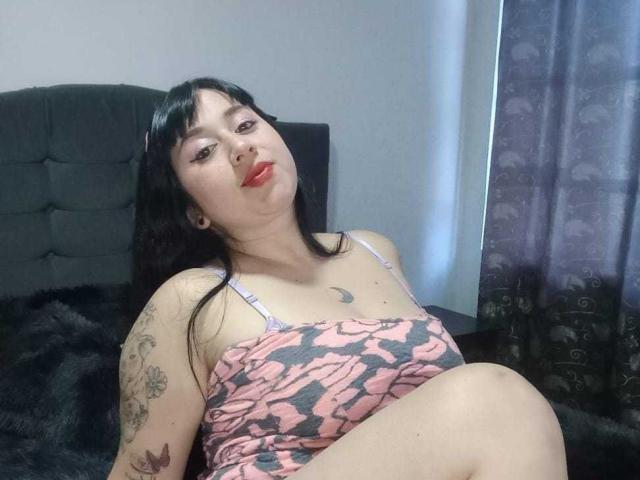 CrystalxSweet with black hair, red lipstick, pink and gray patterned tank top, tattoos on arm, sitting on black bed, looking up, slightly tilted, showing her tits and ass.