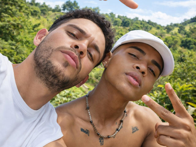 LiorandAxel has a beard, short curly hair, and is shirtless with tattoos, wearing a white cap, pointing with his finger, next to another LiorandAxel with a white shirt.