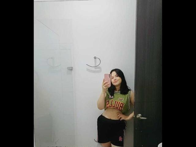 Allanna69 with dark hair, green "FLUAR" crop top, black shorts, holding pink phone, standing in bathroom, smiling, reflecting in mirror.