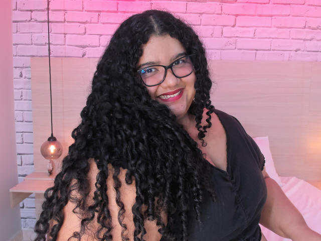 MileyCollinss with long curly black hair, glasses, and red lipstick, smiling at the camera, wearing a black V-neck shirt, showing her large tits and curvy ass.