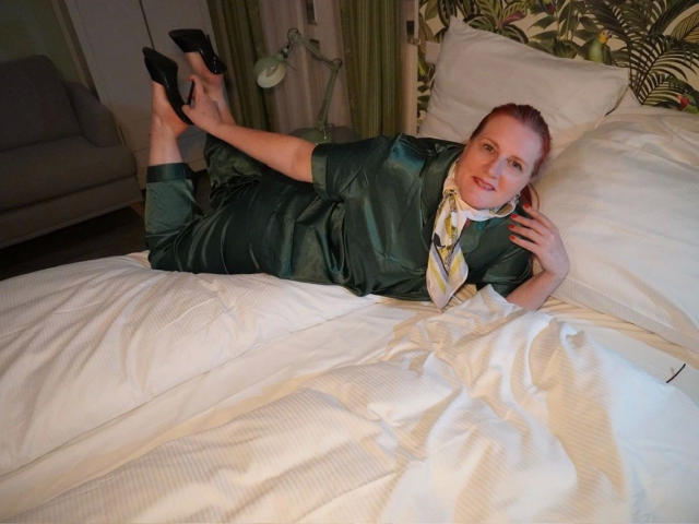 MrsEssen7471-hot, red-haired, lying on white bed, green satin pajamas, black high heels, legs up, white pillow, green patterned headboard, green curtain, smiling, pale skin, neck scarf.