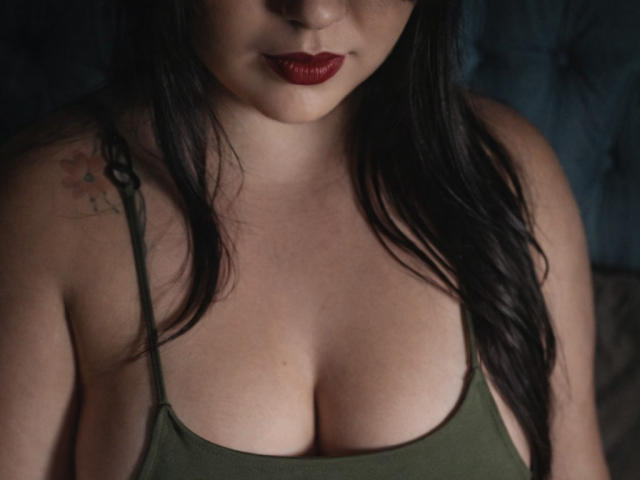 MiaSp's face is partially visible, with red lipstick and dark hair. She's wearing a green tank top, showing her large, soft tits. A small tattoo is on her left shoulder.