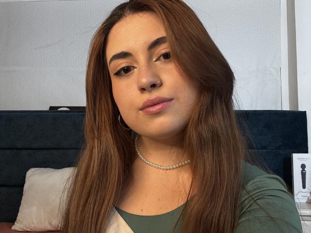 VicluX has long brown hair, subtle makeup, and a pearl necklace, wearing a green shirt, with a small nose ring and a slight, confident smile, looking directly at the camera.