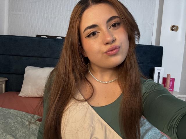 VicluX, com cabelo castanho comprido, blusa verde e branca, um pequeno piercing no nariz, colar de pérolas, olhos verdes e lábios levemente curvados, usando pequenas argolas nos ouvidos, tirando uma selfie..