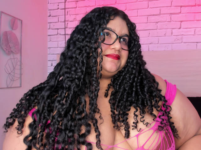 MileyCollinss with curly black hair, glasses, red lipstick, pink bra showing big tits, pink and white brick wall background, slightly smiling, looking at camera.