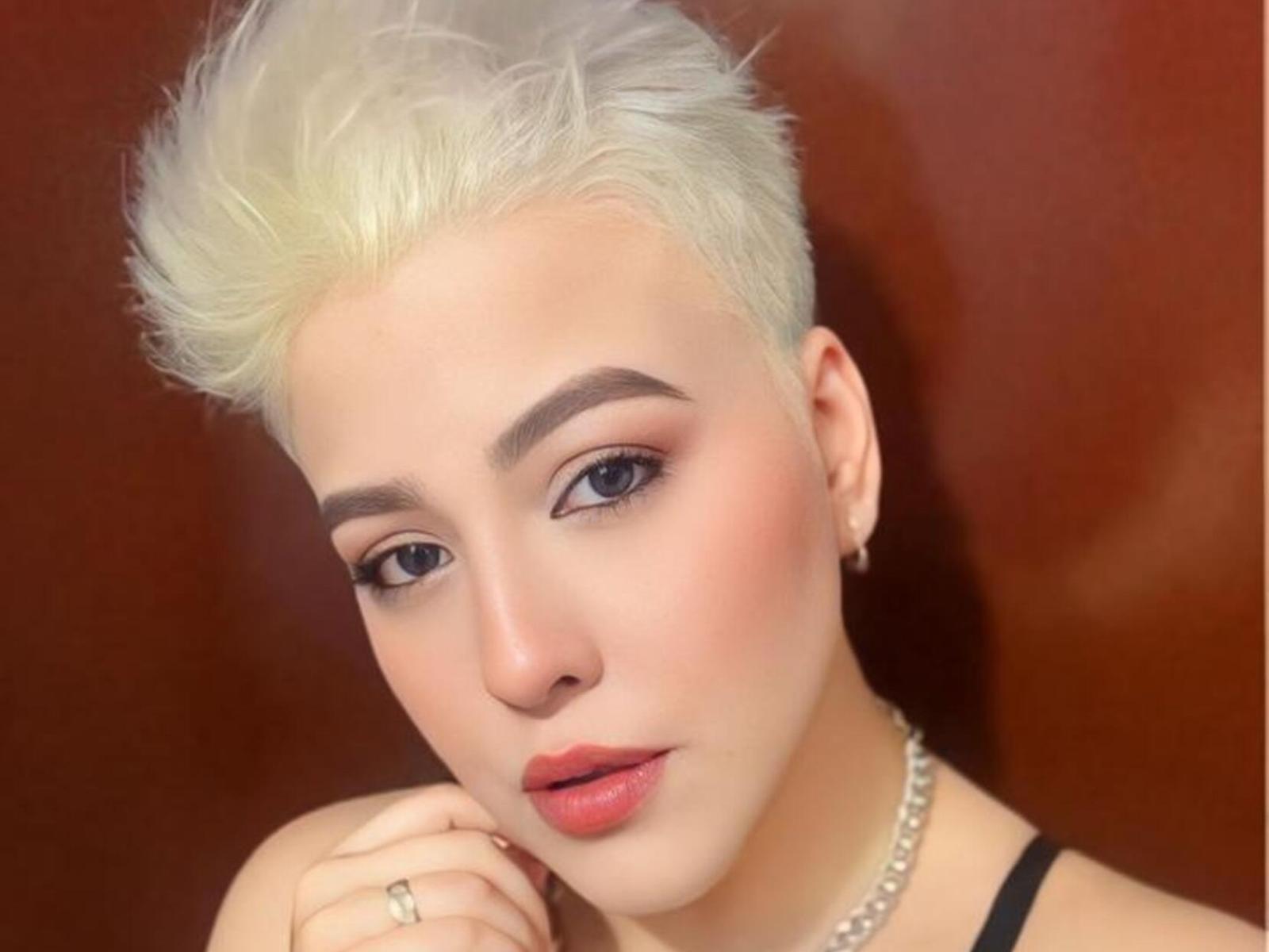 DakotaBailey with short, platinum blonde hair, light skin, thick eyebrows, blue eyes, pink lipstick, and a pearl necklace, holding her chin with a ring on her finger.